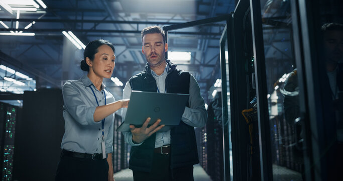 Asian Female Data Analyst Points at Laptop Computer Screen Held by Caucasian Male System Administrator, Colleagues Discussing Server Diagnostics Report and Network Security Scan in Data Center