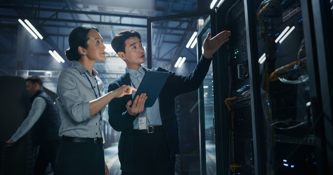 Asian Male Engineer Points at Server Rack Hardware While Holding Tablet Computer, Explaining Cooling System Inspection and AI Infrastructure Audit to Female Coworker in Modern Data Center Facility