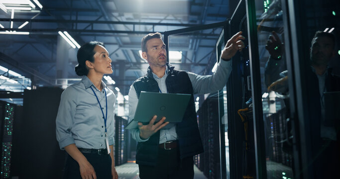 Caucasian Male Technician Holding Laptop Computer Reaches for Hardware Component Inside Open Server Cabinet While Asian Female Engineer Assists, Team Performing AI Infrastructure Repair in Data Center