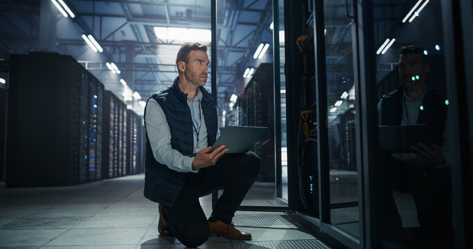 Caucasian Male It Specialist Kneeling Beside Open Server Rack Cabinet While Holding Laptop Computer, Engineer Inspecting Cable Management and Running Network Diagnostics in Modern Data Center