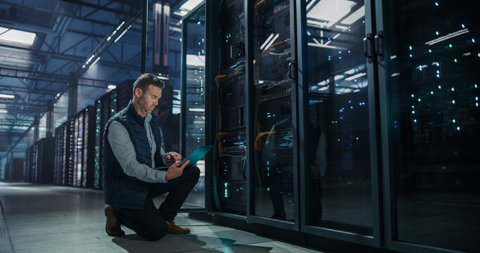 Caucasian Male Network Engineer Kneeling on Floor Beside Server Rack Cabinet, Using Laptop Computer for System Maintenance and Performance Monitoring in Modern AI Data Center Facility
