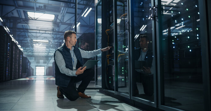 Caucasian Male Data Center Technician Crouching to Reach Into Server Rack Cabinet While Holding Laptop Computer, It Professional Checking Hardware Node and Verifying Storage Array Status