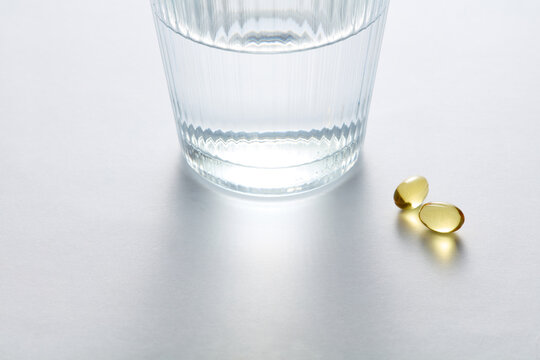 Glass of Clear Water and Two Omega 3 Softgel Capsules on White Surface