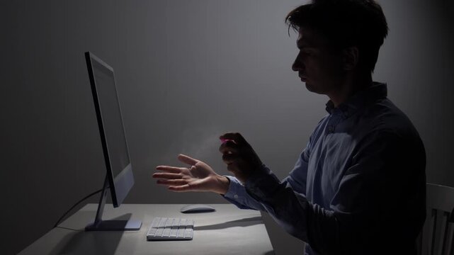 Man using an alcohol spray on his hands for sanitization, prioritizing health and hygiene in a dimly lit home office environment while working on a computer