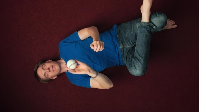 Young man lying on the floor and throwing and catching a white ball in boredom. Slow motion overhead shot with rotation.