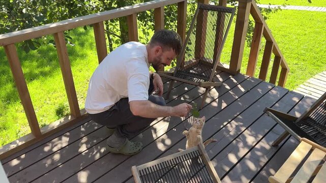 A young man with a beard, who looks like a Scandinavian, is playing with a little red-haired kitten on the porch of a country house. The joy of having a pet, a family pet