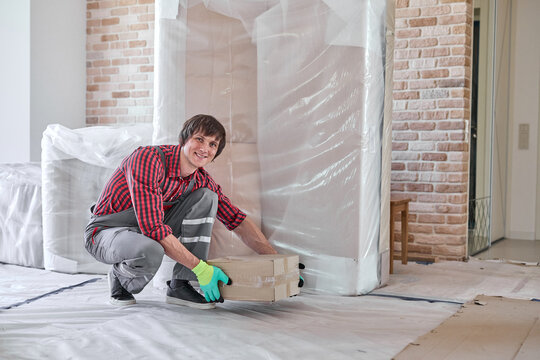 Mover smiling, lifting cardboard box, handling packed furniture in a new apartment during relocation