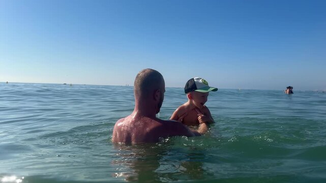 Father plays with his son in the sea, repeatedly lifting him out of the water and letting him jump back into the waves. 4k