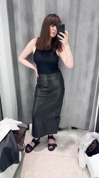 A young woman stands in front of a mirror in a store's fitting room, wearing a black leather skirt and holding her phone; she is trying on clothes and choosing an outfit at a clothing store