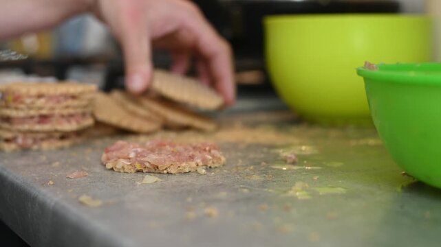 Cook uses metal fork to spread raw minced meat on round waffle sheet. Person prepares homemade waffle meat cutlets inside kitchen. Green plastic bowl stands on messy grey table.