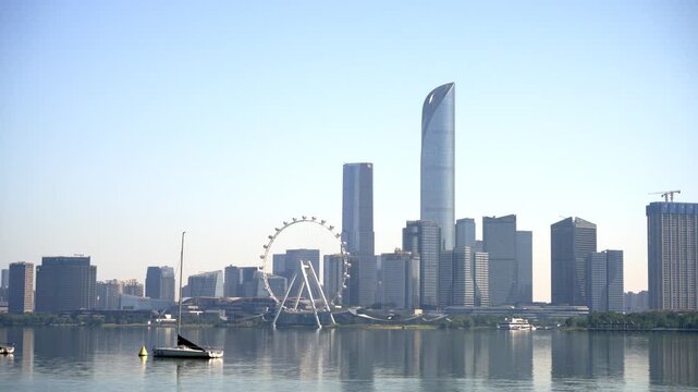 A daytime urban waterfront scene with skyscrapers, a ferris wheel, and building reflections on calm water under a clear sky.