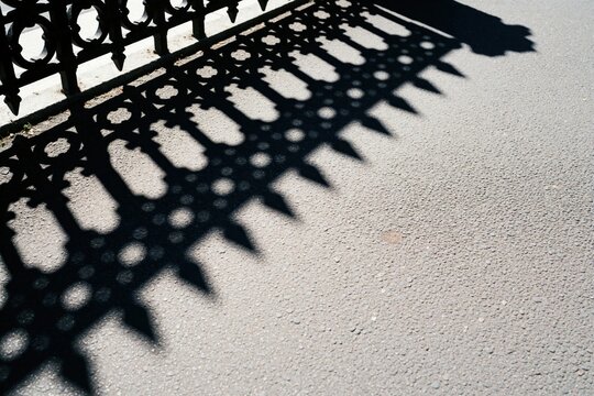 Decorative metal fence casts sharp geometric shadow pattern on grey asphalt ground. Bright sunny daylight creates stark contrast, lines form abstract urban shapes on paved street surface.