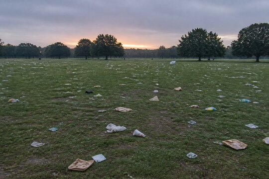 Empty park at dawn with scattered litter and trampled grass, wide shot, flat overcast light, muted desaturated palette, honest documentary look