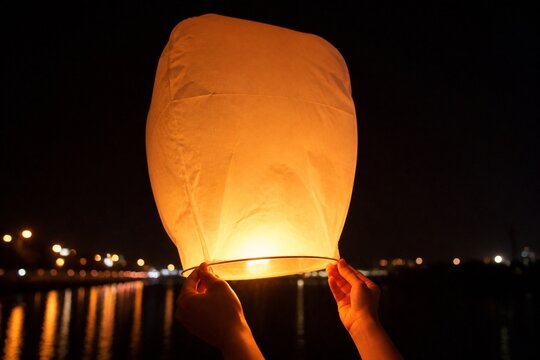 Cropped hands releasing a glowing paper lantern at a riverside festival at night, warm amber glow, plain dark surroundings, no text on the lantern