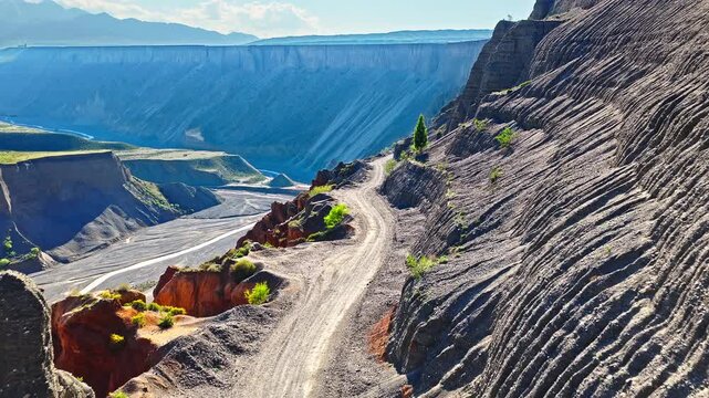 Aerial view of a winding dirt road along a steep eroded canyon cliff with layered geological strata formations in Anjihai Grand Canyon, Xinjiang, China