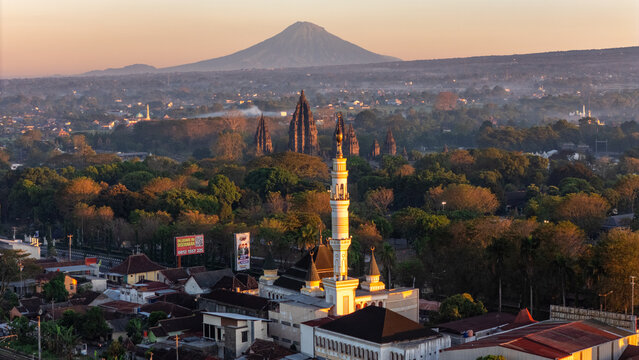 Kecamatan Prambanan, Indonesia - 01 July 2026: Aerial view of Prambanan Temple, a mosque with a tall minaret in the foreground, and a volcanic mountain in the distance at sunset.