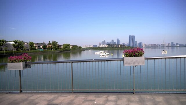 A calm lake scene with city skyline, flower planters on a railing, and boats on the water under clear blue sky.