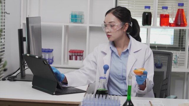 Asian female food scientist wearing lab coat, safety glasses, and gloves examining a fruit sample under a microscope and recording data on a clipboard in a modern microbiology food safety laboratory.
