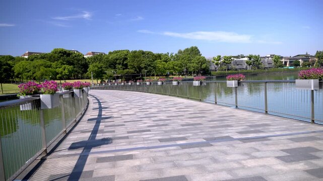 A curved paved walkway runs alongside a calm lake, lined with flower planters and railings, under a clear blue sky with dense trees in the distance.