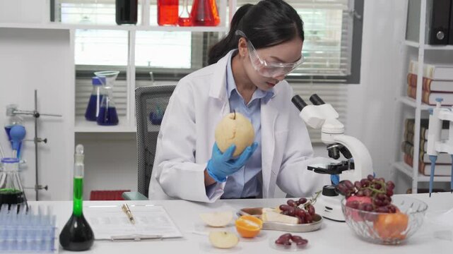 Asian female food scientist wearing lab coat, safety glasses, and gloves examining a fruit sample under a microscope and recording data on a clipboard in a modern microbiology food safety laboratory.