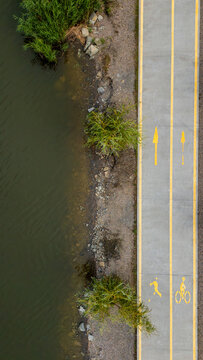 Aerial view presenting a designated concrete bike lane and running path with clear yellow directional arrows and activity symbols. Bordered by a calm river and green vegetation