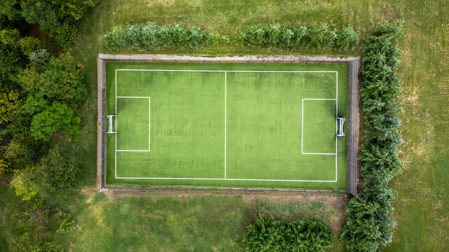 Aerial drone view capturing a vibrant green artificial turf soccer field with clear white markings and goalposts. Enclosed by a protective fence and dense trees