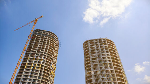Two modern high rise building skeletons are under construction with a large yellow tower crane actively building one, set against a bright blue sky with sparse clouds