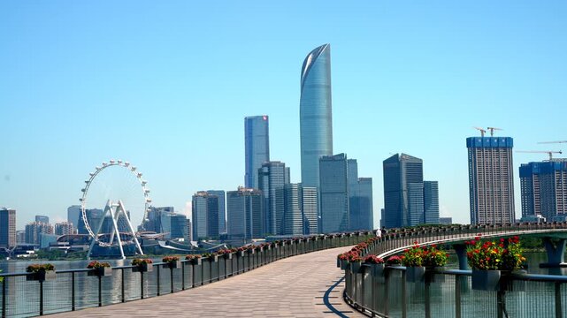 A clear daytime view of a modern urban skyline with skyscrapers, a waterfront path with flower planters, and a ferris wheel in the distance.