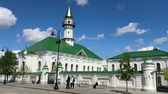 Historic white mosque complex rises beside broad paved plaza, with emerald green roofs, central minaret, arched facade, seated visitor, parked stroller, and pedestrians moving through bright daylight