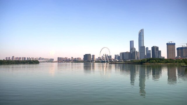 A modern urban skyline with skyscrapers and a ferris wheel is reflected in calm water under a clear blue sky.