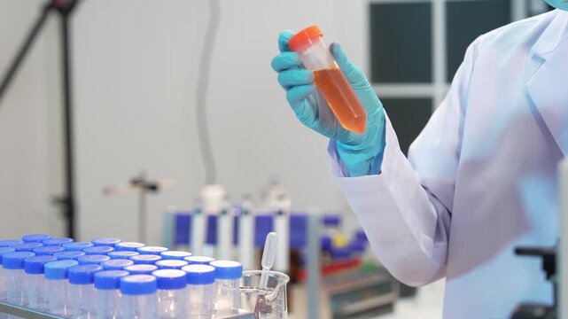 A close-up shot of a female scientist wearing blue gloves pouring various colored liquid chemicals from a bottle into a glass beaker inside a medical or chemical laboratory&mdash;a research concept.