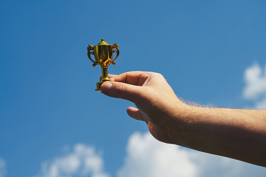 Hand holds and raised small cup trophy, holding cup against clean sky backgrund. Consolation prize award and victory concept.