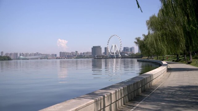 A sunny lakeside walkway with city skyline, ferris wheel, and willow trees along the calm water.