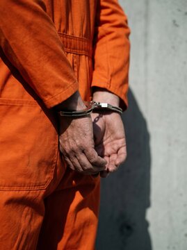 Arrested man in orange prisoner jumpsuit standing with handcuffed hands against concrete wall