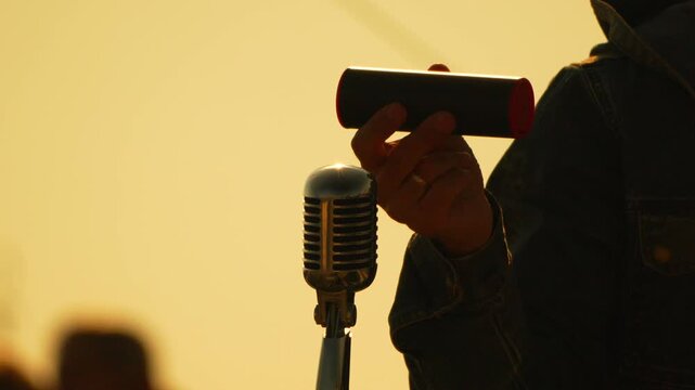 Performer holding portable speaker and silver retro microphone outdoors against a sunset backdrop. Close-up vertical tilt shot.