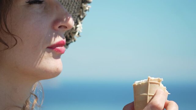 Summer treat enjoyment, a woman wearing a straw hat eats vanilla ice cream near the sparkling sea. Static profile close-up shot.