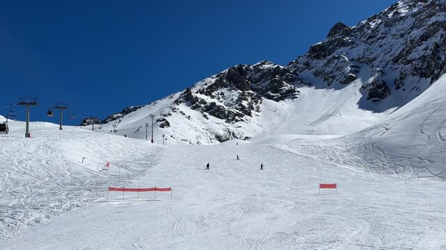 Wide mountain ski run with groomed snow, small skiers moving downhill, chairlift towers, rugged snowy peaks, red markers, and clear blue sky in bright winter light across open alpine terrain