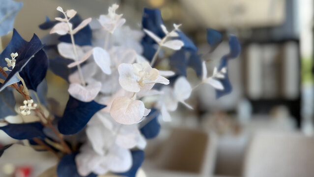 Close-up of decorative blue and white artificial floral arrangement in a contemporary interior, with soft natural light and blurred modern living space in the background.