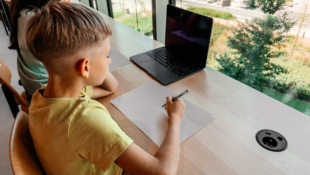 Young schoolboy doing homework with a laptop at a desk while studying online. Digital education, remote learning, schoolwork and home study concept.