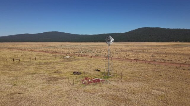 Drone executes orbiting approach toward active windmill on golden rangeland with mountain backdrop, Lassen County, California.