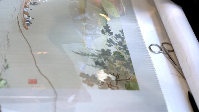 An artisan works on delicate hand embroidery, using needle and thread on translucent silk fabric, with scissors placed nearby.