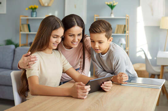 Happy mother sitting with daughter and son at home, watching something on tablet together. Smiling parent and children enjoying family time, engaging with entertaining content online and having fun.