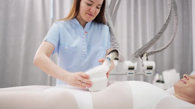 Female client wearing a specialized white treatment suit receives professional non-invasive body contouring treatment with aesthetic equipment at a modern beauty clinic.