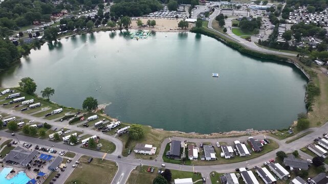 An aerial drone shot orbits above Sherkston Quarry, highlighting the floating inflatable water park obstacle course, the sandy beachfront, and the surrounding holiday RV campgrounds