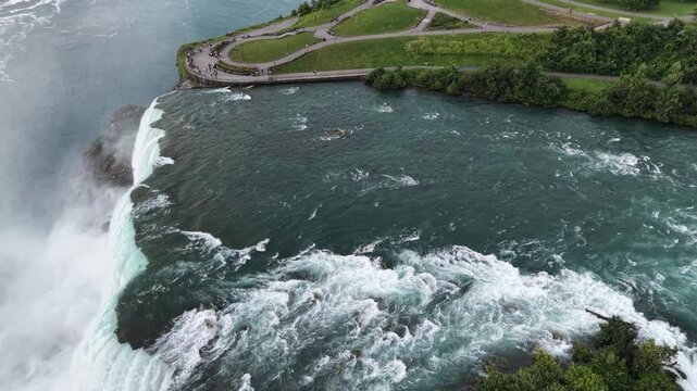 High-angle aerial tracking shot following along the curved green water crest, rushing white water rapids, and green rocky islets at the brink of the Horseshoe Falls, Canada
