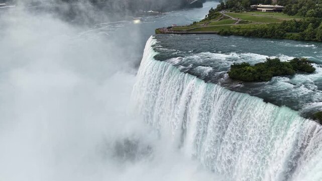 Wide aerial tracking shot moving forward over a thundering vertical water plunge, green rocky islands, and massive plumes of white mist at the crest of the Horseshoe Falls, Canada