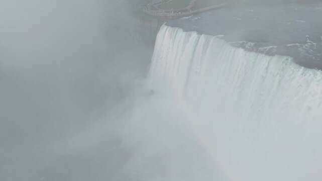 An aerial drone shot orbits right over the thundering brink of the Horseshoe Falls, revealing the cliffside observation deck and a distant tour boat navigating the misty river below