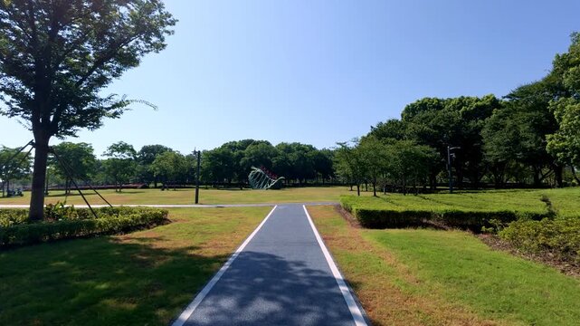 A bright sunny day view of a paved park path surrounded by green grass and trees, with open landscape in the distance.