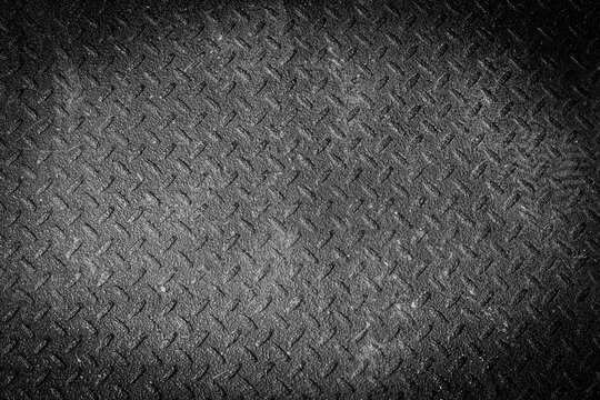 Close-up abstract texture of a dark black painted diamond plate surface with a repeating raised tread pattern and rugged industrial appearance. High-contrast lighting emphasizes the textured surface.