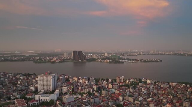 Drone view of dense Hanoi neighborhoods, West Lake, modern development, and a distant city skyline beneath a colorful evening sky.